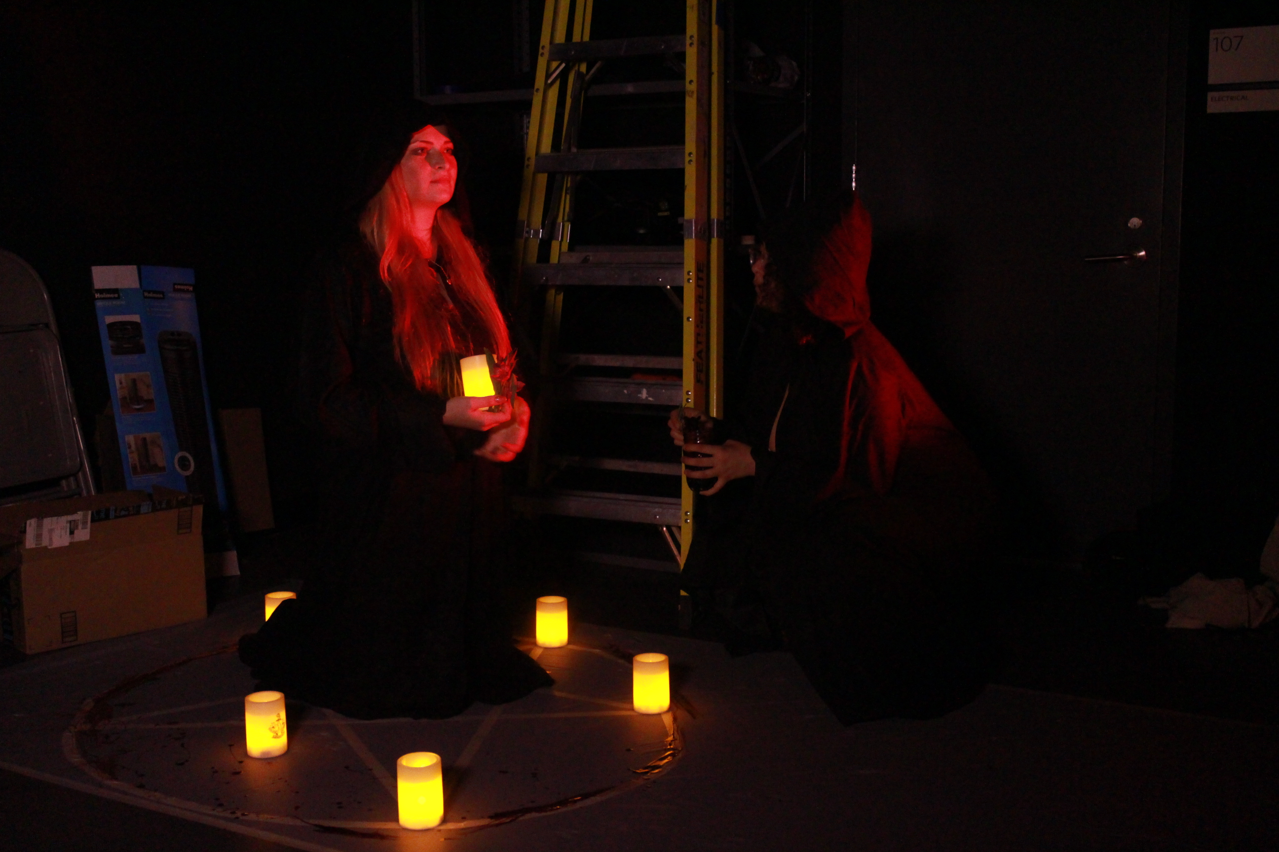 The candles on the pentagram are lit up in the darkness, and two student performers are in dark hooded robes holding candles in their hands.