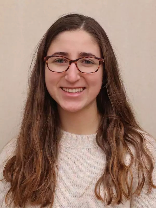 a white young woman with brown hair and glasses, wearing a beige shirt