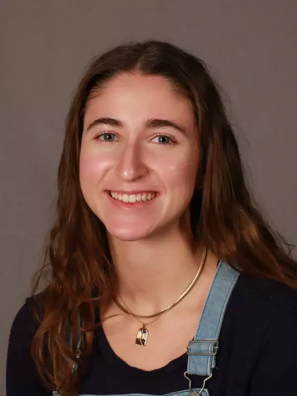 A white young woman with brown hair and blue/green eyes and wearing a blue shirt and denim overalls